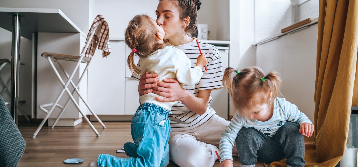 Äiti ja kaksi lasta istuvat lattialla ruokapöydän edessä. Äiti antaa toiselle lapsista pusua.