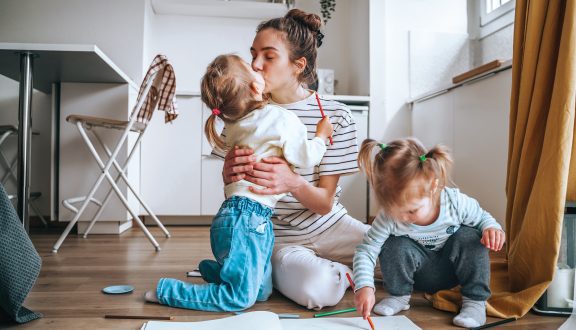 Äiti ja kaksi lasta istuvat lattialla ruokapöydän edessä. Äiti antaa toiselle lapsista pusua.