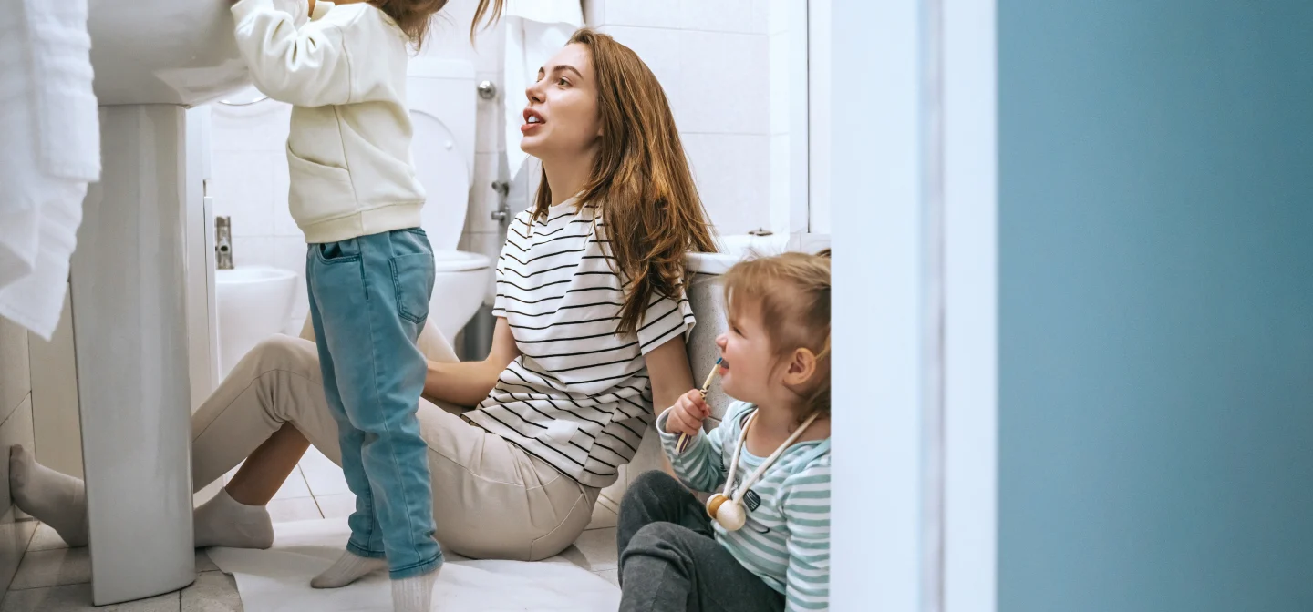 Nainen istuu vessan lattialla kahden lapsen kanssa. Toinen lapsista seisoo ja kurkottaa lavuaarin yli.