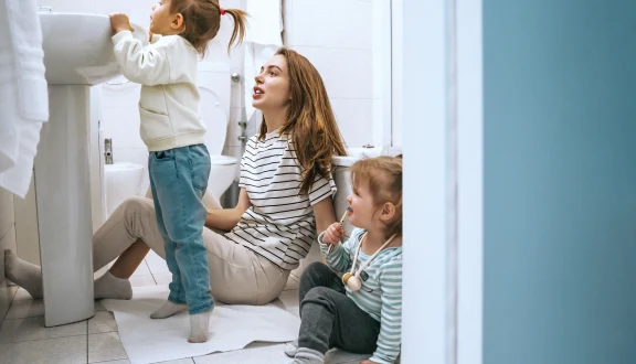 Nainen istuu vessan lattialla kahden lapsen kanssa. Toinen lapsista seisoo ja kurkottaa lavuaarin yli.