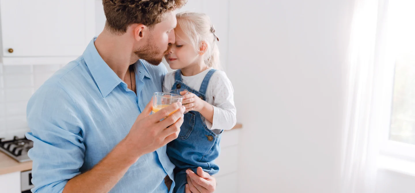 Isä seisoo lapsi sylissä ja lasi toisessa kädessä valoisassa huoneessa.