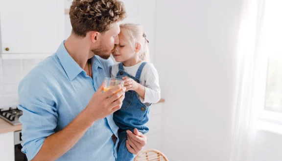 Isä seisoo lapsi sylissä ja lasi toisessa kädessä valoisassa huoneessa.