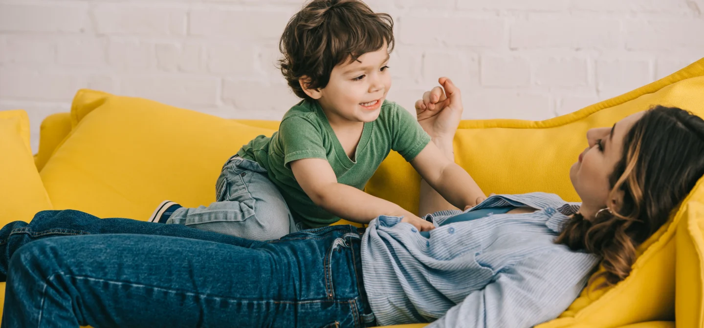 Äiti makaa keltaisella sohvalla. Lapsi makaa hänen mahansa päällä irvistäen.