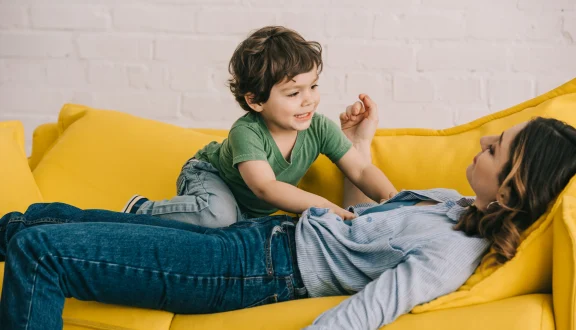 Äiti makaa keltaisella sohvalla. Lapsi makaa hänen mahansa päällä irvistäen.