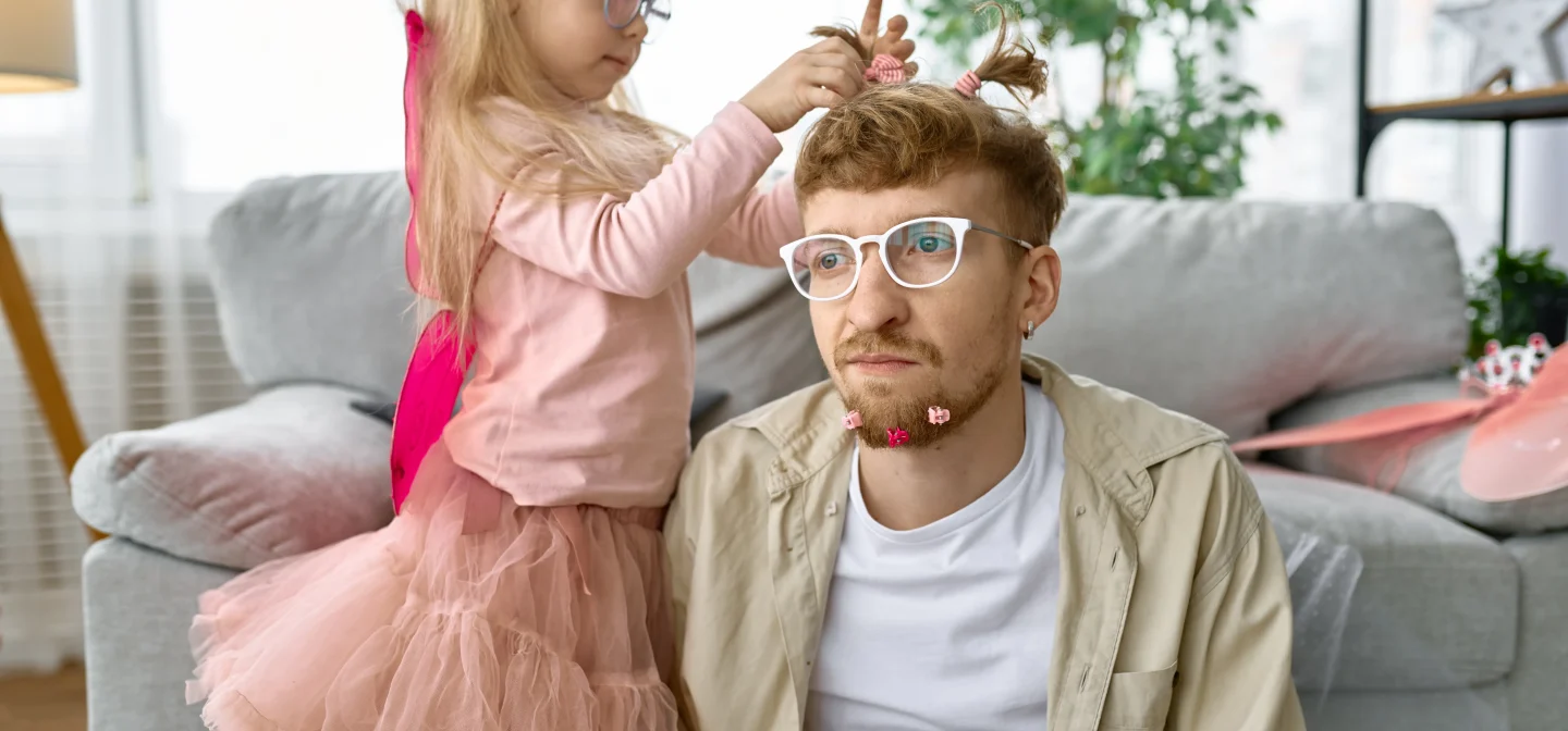 Isä istuu sohvan edessä lattialla, samalla kun tytär prinsessa-asussa letittää hänen tukkaansa.