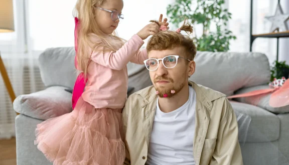 Isä istuu sohvan edessä lattialla, samalla kun tytär prinsessa-asussa letittää hänen tukkaansa.
